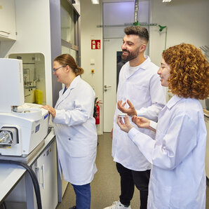 Studierende im Laborkittel lächeln und unterhalten sich neben einem Laborgerät.