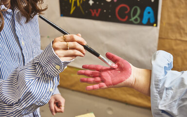 Partizipation Ein studierende malt mit einem Pinsel die Hand einer anderen Person mit roter Farbe an