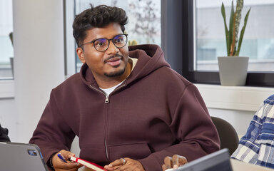 Ein Studierender sitzt an einem Tisch mit Papieren und Laptop ins Gespräch vertieft