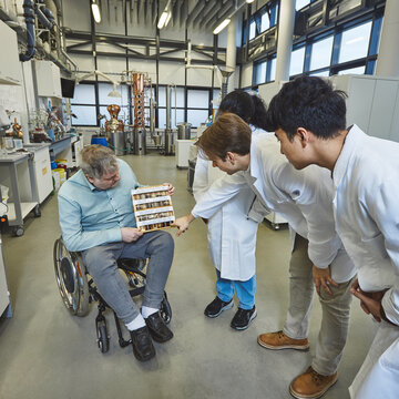 Untersuchung von Bodenproben - Ein Dozent im Rollstuhl zeigt drei Studierenden in Kitteln ein Gestell mit verschiedenen Boden- und Materialproben In einem hellen Labor sitzt ein Mann im Rollstuhl und hält ein hölzernes Präsentationsgestell mit vielen kleinen Probenröhrchen auf dem Schoß. Drei Studierende in weißen Laborkitteln beugen sich neugierig zu ihm herunter und betrachten die Proben.