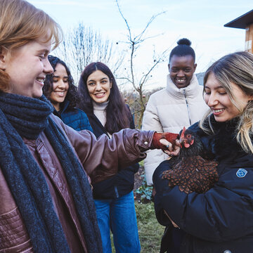 Sechs Studierende stehen im Halbkreis um eine Studentin, die ein Huhn auf dem Arm hält. Eine Studentin streichelt das Huhn.