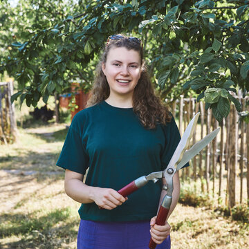 Eine junge Studentin steht an einem sonnigen Tag in einem grünen Garten unter einem Baum. Sie lächelt in die Kamera und hält eine große Astschere in beiden Händen vor dem Körper.