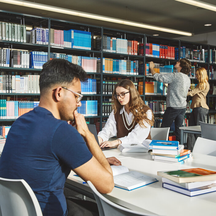 Fünf Studierende sitzen an Arbeitsplätzen oder stehen an den Bücherregalen im Lesesaal der Bibliothek Kamp-Lintfort