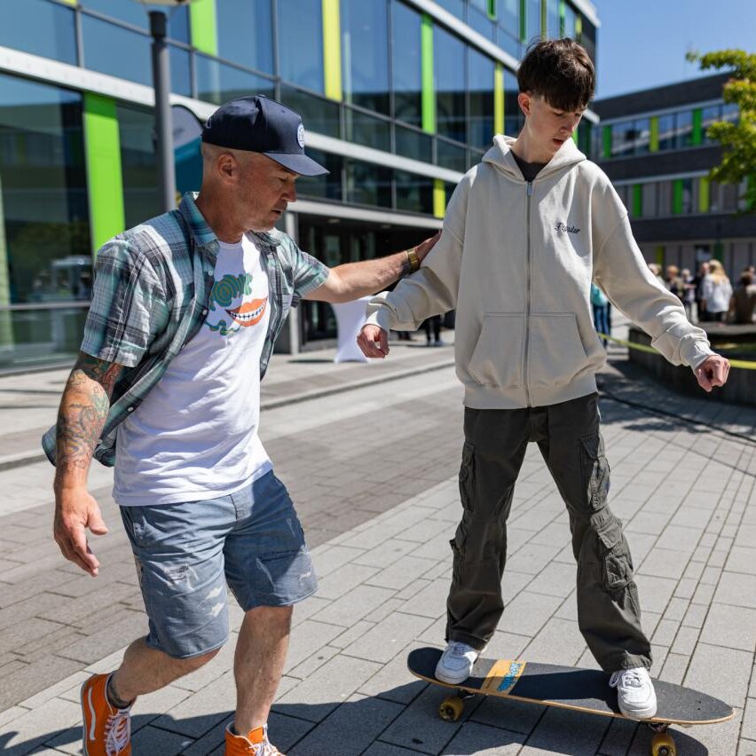 Studierender übt Skate Board fahren auf dem Campus mit Anleitung