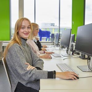 Studentin sitzt lächelnd an einem Computerarbeitsplatz in einem hellen Computerlabor.