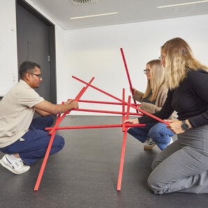 Three students are kneeling on the floor, building a structure out of red rods.