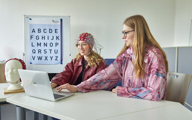 Zwei Studentinnen arbeiten gemeinsam am Laptop; eine von ihnen trägt eine EEG-Kappe mit Elektroden zur Messung der Gehirnaktivität