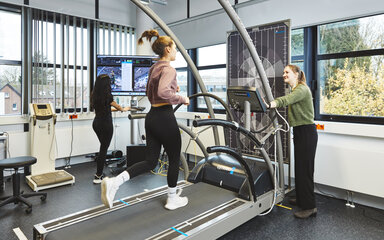 Eine junge Frau joggt auf einem großen Laufband im Labor. Rechts von ihr steht eine lächelnde Studentin und bedient das Gerät. Im Hintergrund arbeitet eine weitere Studentin aufmerksam an einem Monitor, der Videobilder und Daten der Läuferin anzeigt.