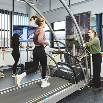 Eine junge Frau joggt auf einem großen Laufband im Labor. Rechts von ihr steht eine lächelnde Studentin und bedient das Gerät. Im Hintergrund arbeitet eine weitere Studentin aufmerksam an einem Monitor, der Videobilder und Daten der Läuferin anzeigt.