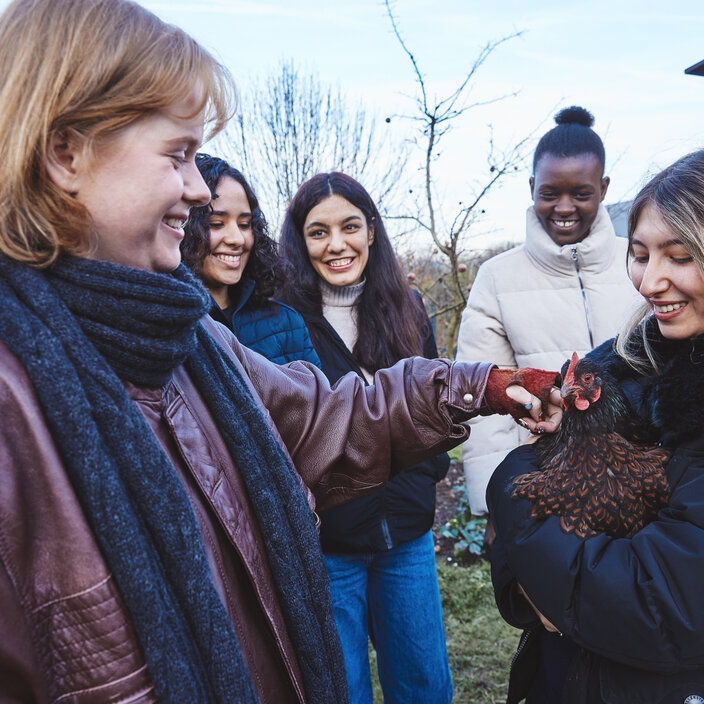 Gruppe um das Huhn - Agrarwirtschaft hautnah erleben Eine lachende Studentin hält ein Huhn auf dem Arm. Um sie herum stehen mehrere Kommilitoninnen und Kommilitonen, die neugierig zuschauen; eine Person streichelt dem Huhn vorsichtig über den Kopf.