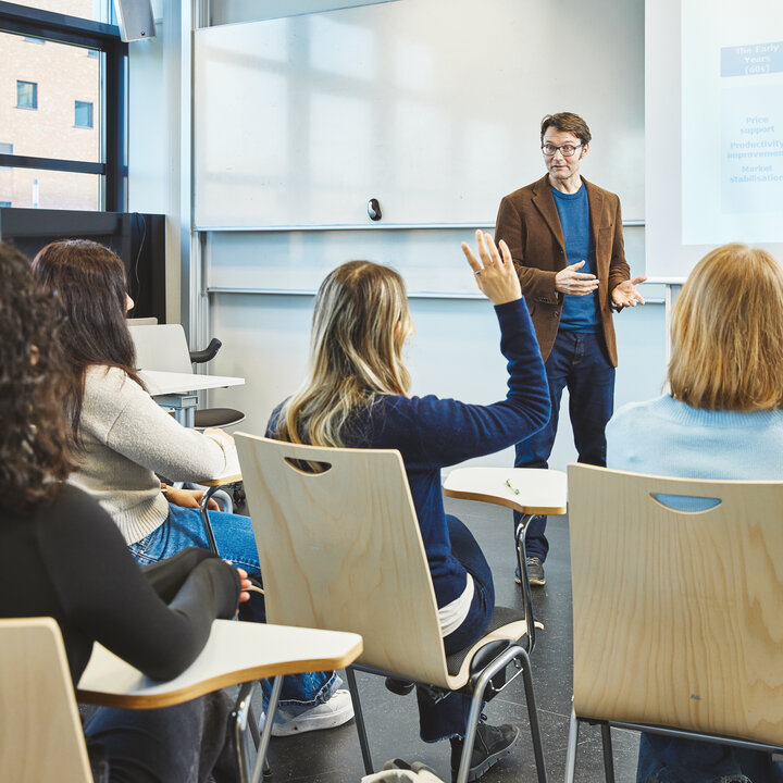 A lecturer is standing in a brightly lit lecture theatre in front of a presentation slide, speaking to the students. A female student in the foreground raises her hand to ask a question.