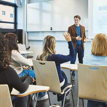 Dozent vor der Klasse - Interaktive Vorlesung im Agribusiness Ein Dozent steht in einem hellen Hörsaal vor einer Präsentationsfolie und spricht zu den Studierenden. Eine Studentin im Vordergrund meldet sich mit gehobener Hand.