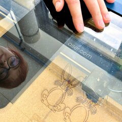 A young woman with reddish hair and glasses is leaning over a laser cutter. In the background, two cut-out rabbits can be seen.