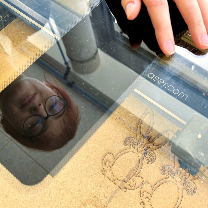 A young woman with reddish hair and glasses is leaning over a laser cutter. In the background, two cut-out rabbits can be seen.
