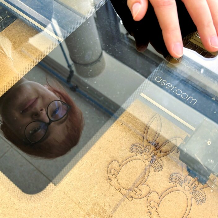 Eine junge Frau mit rötlichen Haaren und Brille beugt sich über einen Lasercutter. Im Hintergrund sind zwei ausgeschnittene Hasen zu sehen.
