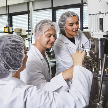 Detailarbeit an lebensmitteltechnologischen Anlagen im Team Drei Studierende in Laborkitteln und Haarnetzen arbeiten konzentriert zusammen an den Ventilen und Armaturen eines großen Edelstahl-Bioreaktors im Technikum.