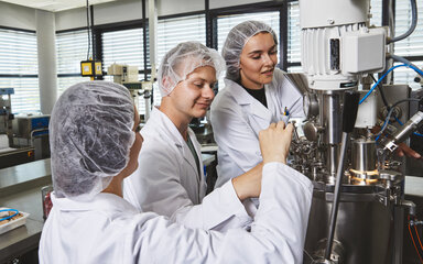 Detailarbeit an lebensmitteltechnologischen Anlagen im Team Drei Studierende in Laborkitteln und Haarnetzen arbeiten konzentriert zusammen an den Ventilen und Armaturen eines großen Edelstahl-Bioreaktors im Technikum.
