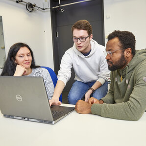 Drei Studierende sitzen gemeinsam vor einem Laptop und besprechen ihre Arbeit.