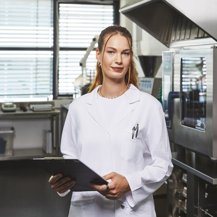 Eine junge Studentin im weißen Laborkittel hält ein schwarzes Klemmbrett in den Händen und blickt selbstbewusst in die Kamera. Im Hintergrund befindet sich ein Labor mit industriellen Edelstahlöfen und Lüftungsanlagen.