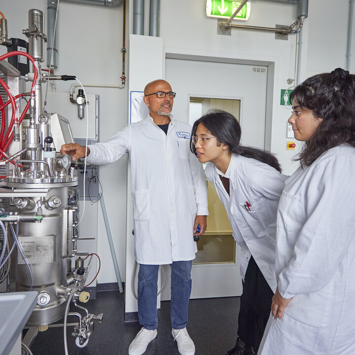A photo of a lecturer and two students in white lab coats looking together at the components of a large stainless-steel bioreactor with numerous tubes.