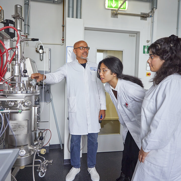 Foto eines Dozenten und zweier Studierender in weißen Kitteln, die gemeinsam auf die Instrumente eines großen Edelstahl-Bioreaktors mit zahlreichen Schläuchen schauen.