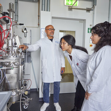 Bioengineering, B.Sc. Foto eines Dozenten und zweier Studierender in weißen Kitteln, die gemeinsam auf die Instrumente eines großen Edelstahl-Bioreaktors mit zahlreichen Schläuchen schauen.