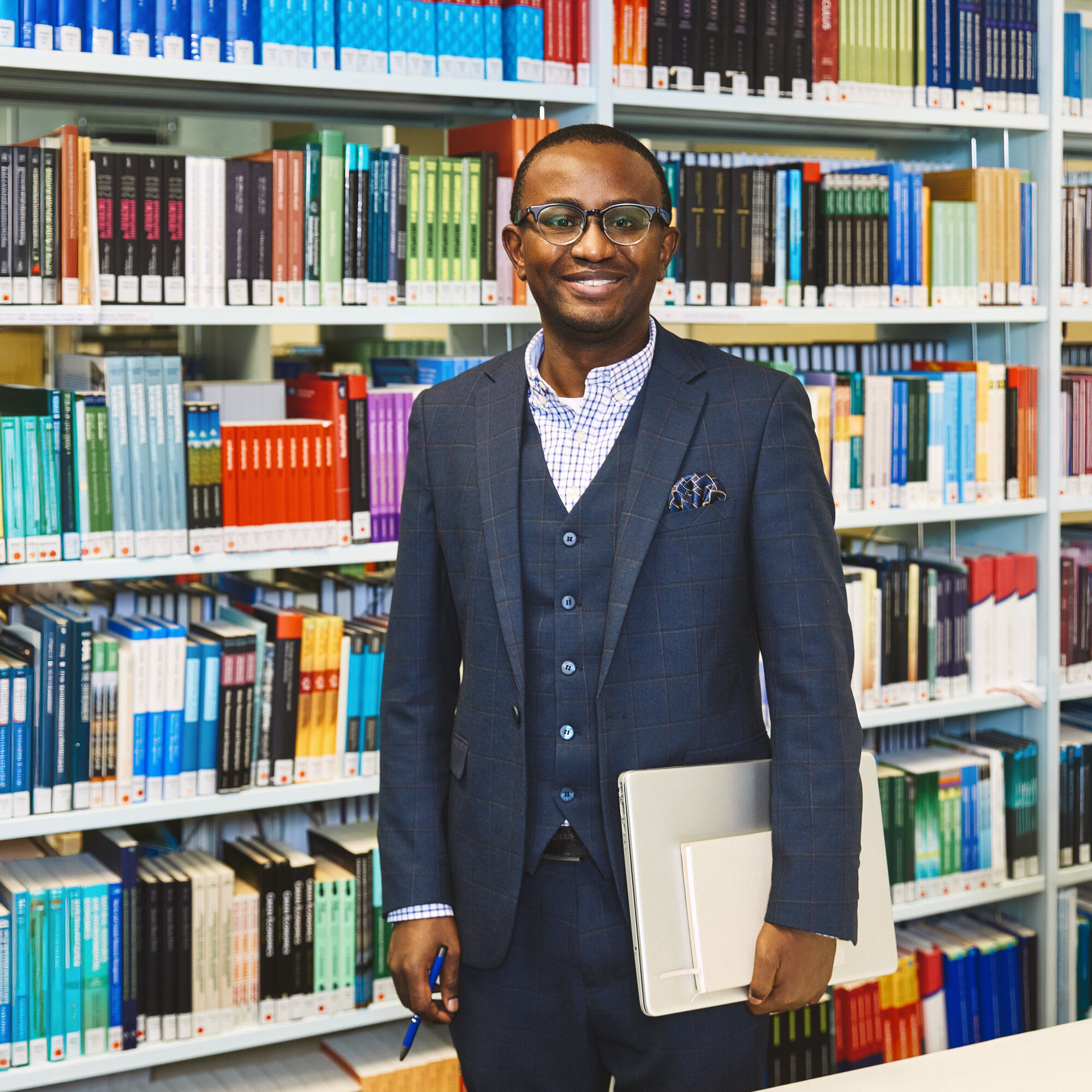 A student with a laptop is standing in front of a bookshelf in Kleve Library, smiling at the camera