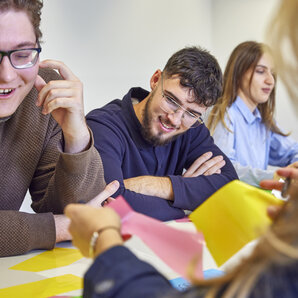 Studierende sitzen an einem Tisch voller bunter Haftnotizen und lächeln.