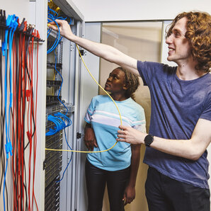 Zwei Studierende stehen im Elektroniklabor vor einer Wand mit zahlreichen Kabeln.