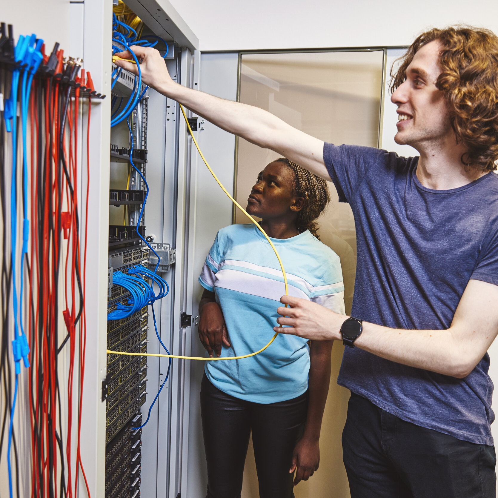 Zwei Studierende stehen im Elektroniklabor vor einer Wand mit zahlreichen Kabeln.