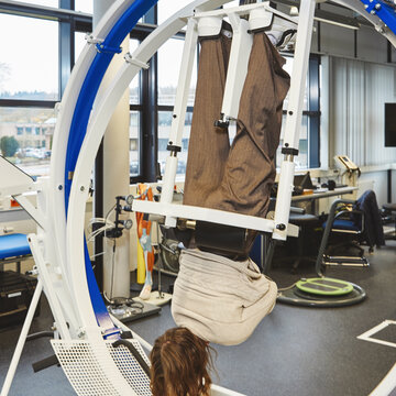 Eine Person liegt in einer großen, ringförmigen 3D-Trainingsanlage. Ihr Körper ist in einem weißen Gestell fixiert, sodass sie kopfüber hängt, während sie das Gerät für eine Untersuchung nutzt.