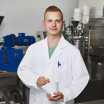 Student bereitet Lebensmittelproben mit Mörser und Pistill vor Ein junger Student im weißen Laborkittel steht an einem Arbeitstisch aus Edelstahl und reibt mit einem Pistill in einem weißen Porzellanmörser. Im Hintergrund sind verschiedene blaue und silberne Laborgeräte zu erkennen.