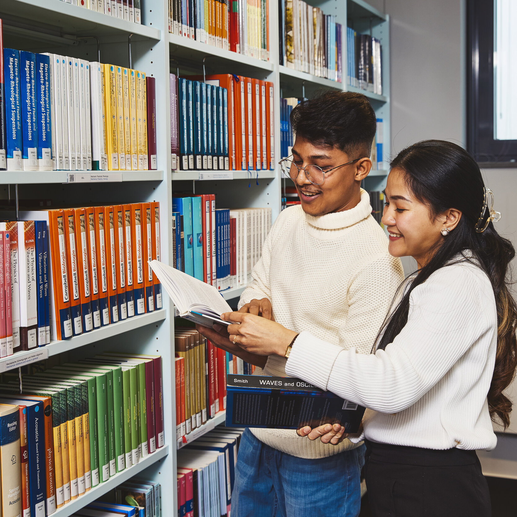 Zwei Studierende stehen vor einem Bücherregal der Bibliothek Kleve und sehen sich gemeinsam Bücher an