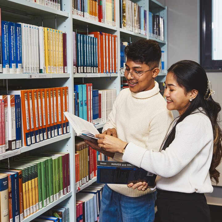 Zwei Studierende stehen vor einem Bücherregal der Bibliothek Kleve und sehen sich gemeinsam Bücher an
