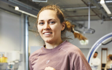 Running for empirical research – A young woman is jogging cheerfully on a treadmill in the health laboratory A young woman with her hair tied back in a plait is jogging on a treadmill in sportswear, smiling cheerfully at the camera. In the blurred background, medical equipment can be seen in a brightly lit laboratory.