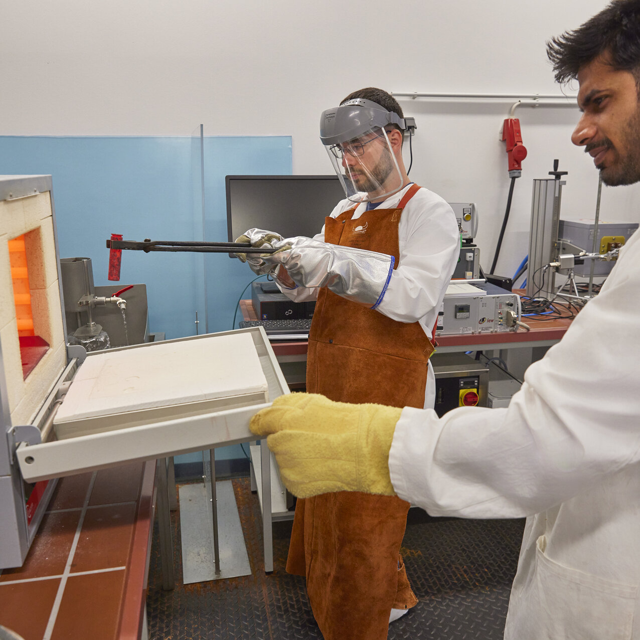 Ein Studierender in Schutzkleidung steht an einem geöffneten Hochtemperaturofen und hält in seinen Händen eine Zange mit einer Materialprobe.