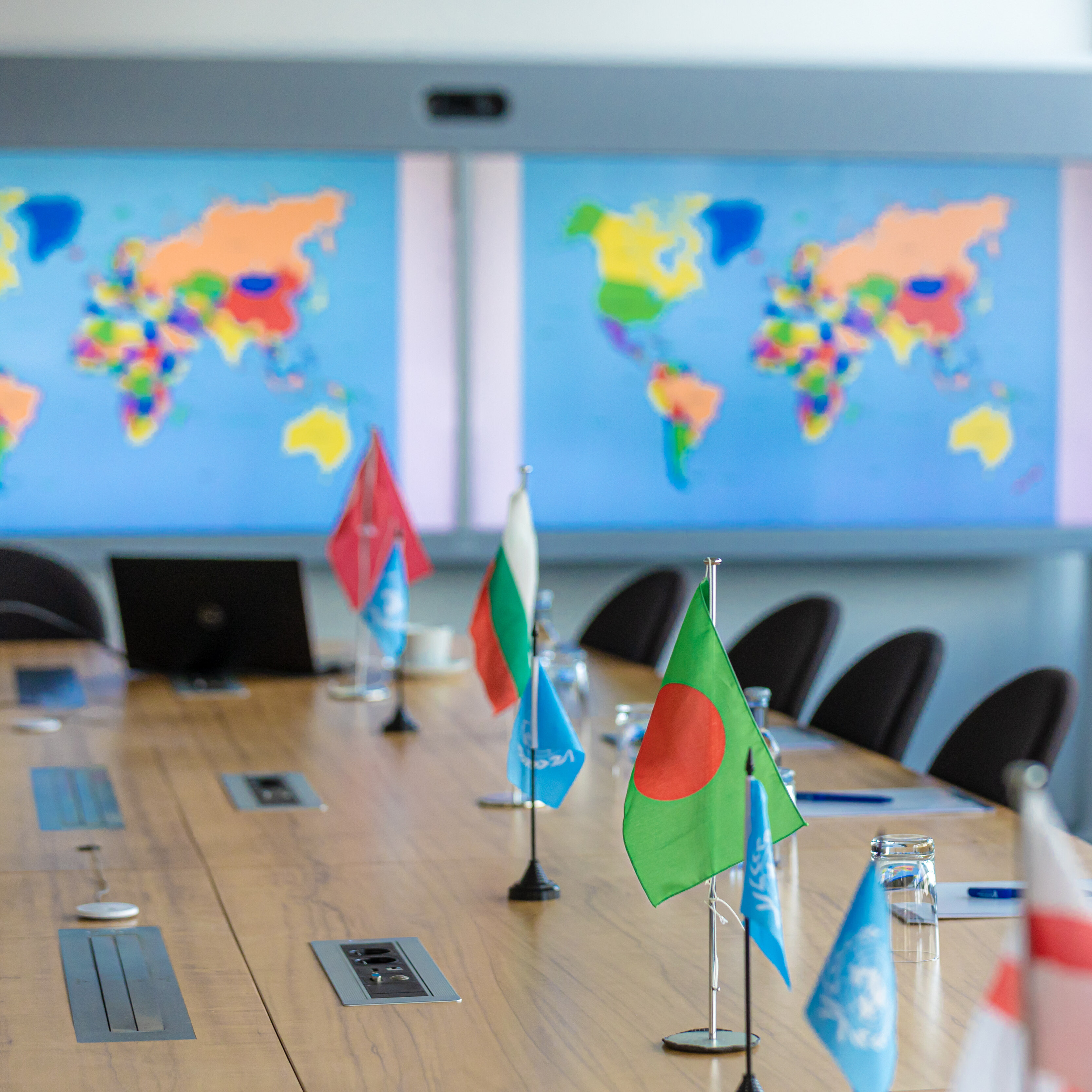 Ein Konferenztisch mit Tischflaggen. Im Hintergrund auf einem großen Bildschrim eine Weltkarte und rechts das Bannder der Studierendeniniative KleMUN