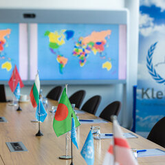 A conference table with table flags. In the background, a world map is displayed on a large screen, and to the right is the banner of the student initiative KleMUN.