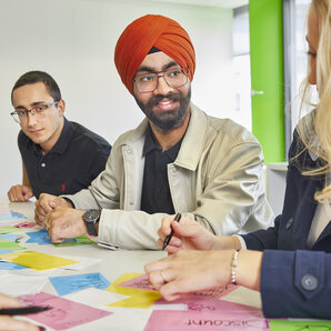 Studierende sitzen an einem Tisch voller bunter Haftnotizen, schreiben auf Notizen und unterhalten sich.