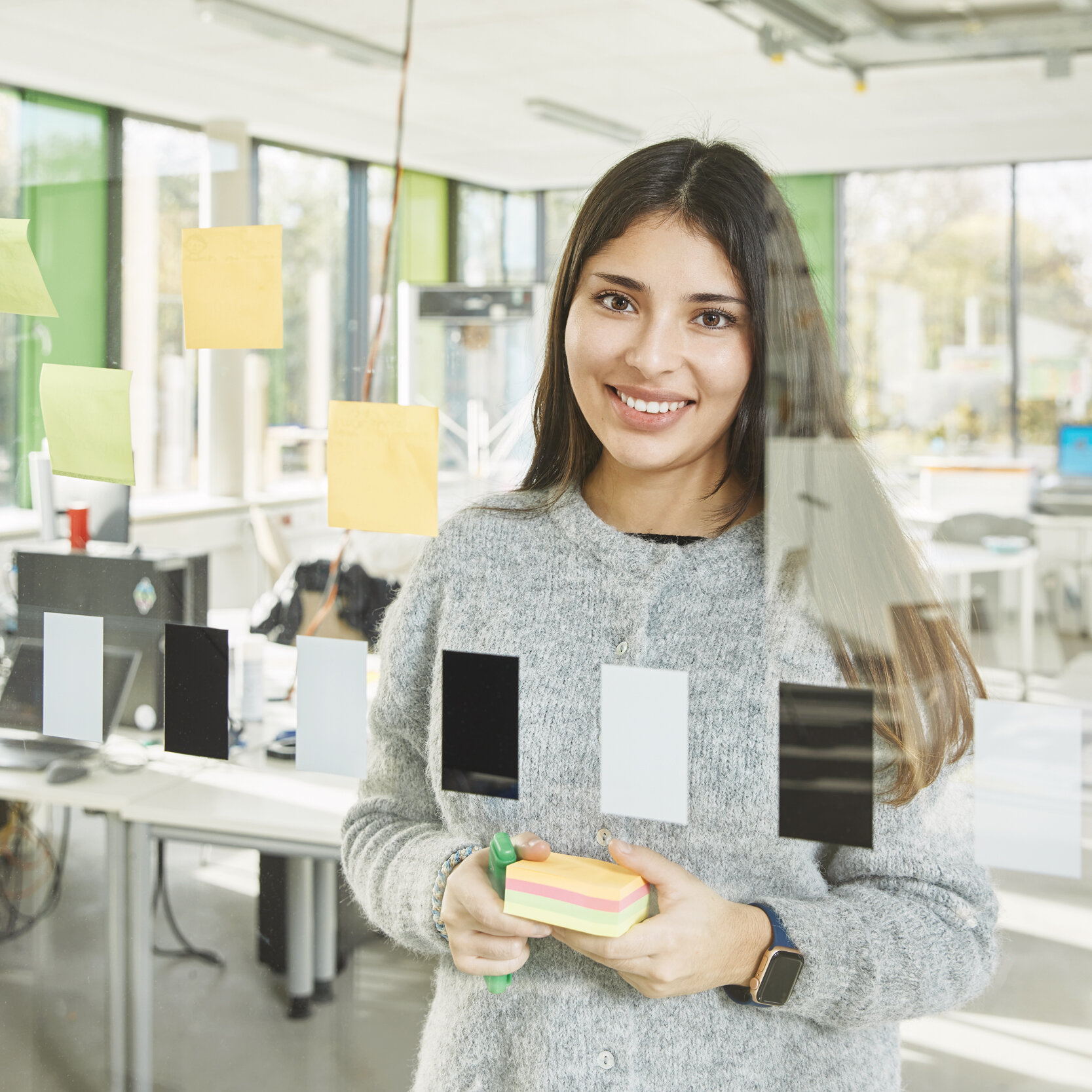 Eine junge Studentin steht hinter einer Glasscheibe, auf der Klebezettel angebracht sind. Sie lächelt in die Kamera.