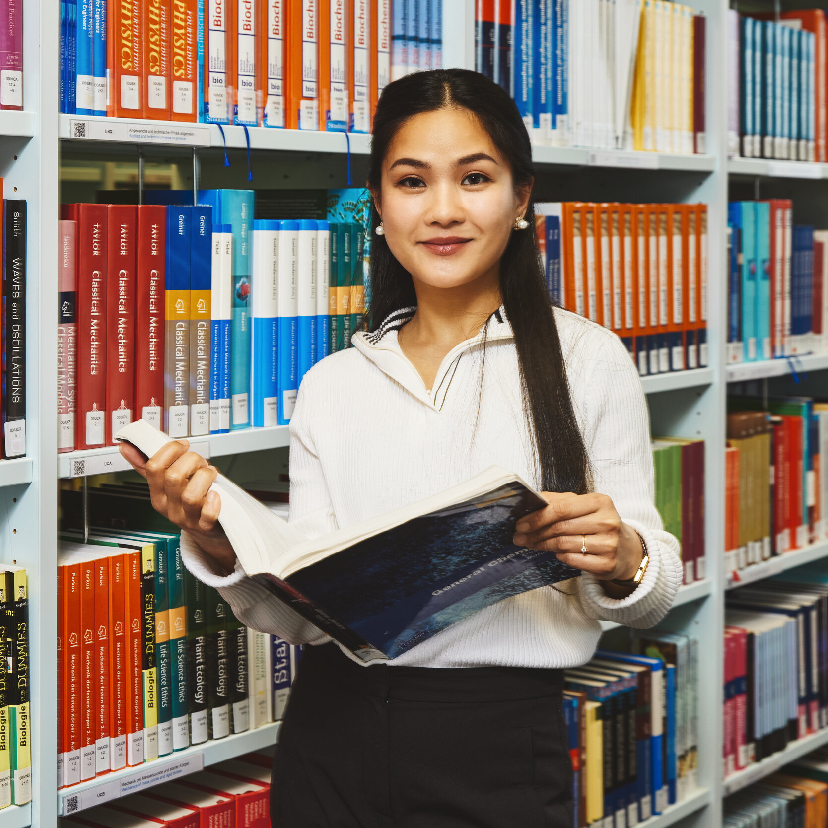 Eine Studentin steht mit einem Buch vor einem Bücherregal in der Bibliothek Kleve und lächelt in die Kamera