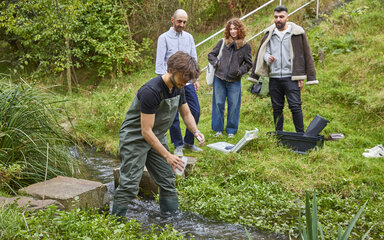 Deine Zukunft: grün & gefragt Student in Wathose entnimmt eine Wasserprobe aus einem Bach, während drei weitere Studierende zusehen.