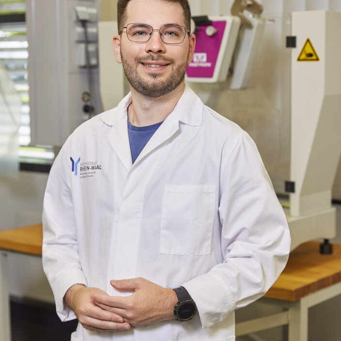 Fabio Ein Studierender steht im weißen Laborkittel in einem Chemielabor und schaut lächelnd in die Kamera.