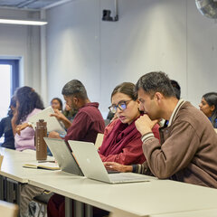 Eine Gruppe Studierender befindet sich in einem Seminarraum. Im Fokus des Bildes sitzen zwei Studierende an einem Tisch und schauen auf einen Laptopbildschirm.