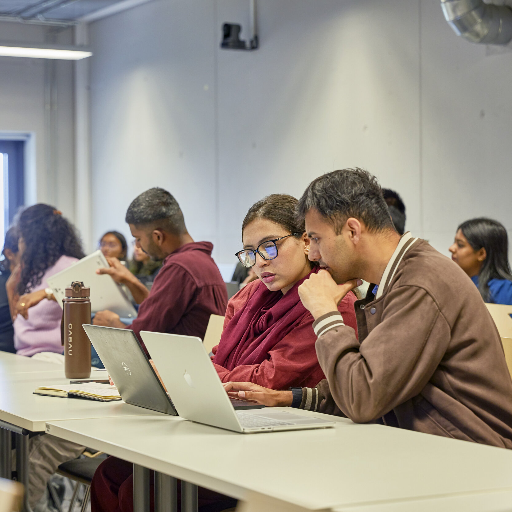 Eine Gruppe Studierender befindet sich in einem Seminarraum. Im Fokus des Bildes sitzen zwei Studierende an einem Tisch und schauen auf einen Laptopbildschirm.