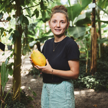 Eine junge Studentin steht in einem dicht bewachsenen tropischen Gewächshaus unter großen Bananenstauden. Sie hält lächelnd eine große, gelbe Papaya in beiden Händen vor dem Bauch.