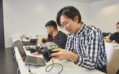 Zwei Welten, ein Studium Studierende sitzen an Laptops, ein Student arbeitet an einem elektronischen Gerät.