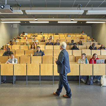 Das Bild zeigt einen kleinen menschengefüllten modernen Hörsaal in dem ein Lehrender vor den Studierenden steht und eine Vorlesung hält.