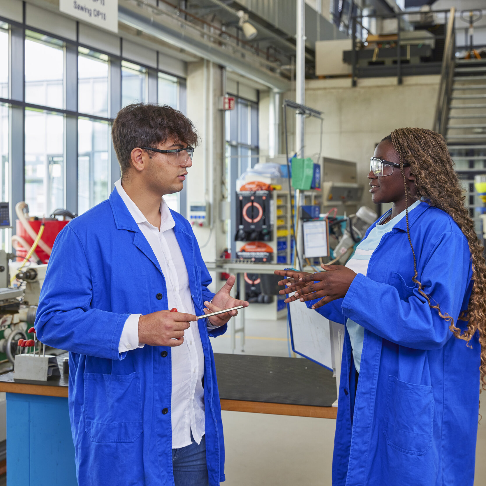 Zwei Studierende stehen in Schutzkleidung und mit Schutzbrille in der Technologiehalle der Fakultät. Sie sind einander zugewandt und unterhalten sich.