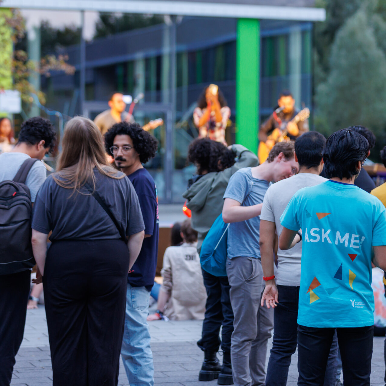 In front of a stage on Kamp-Lintfort campus, a large number of students are seen with their backs to the camera, watching the Music Night. A tutor’s T-shirt reads ‘Ask Me’. 